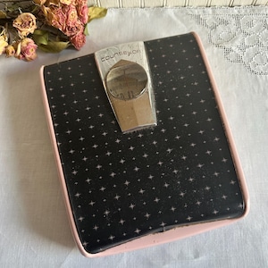 May include: Vintage black and pink bathroom scale with a starburst pattern. The scale has a silver-colored dial and the word "COUNSELOR" is printed on the top. The scale has a pink border.