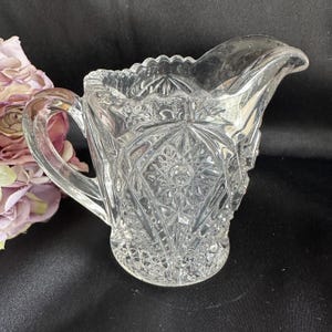 May include: Clear glass creamer with a handle and spout. The creamer features a detailed, cut-glass design with diamond and starburst patterns. The rim has a scalloped edge. The creamer is likely vintage or antique.