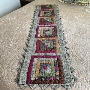 May include: A rectangular quilted table runner featuring a series of five square log cabin blocks. The blocks are constructed from various patterned fabrics in shades of burgundy, olive green, brown, and tan, bordered by a speckled gray fabric.