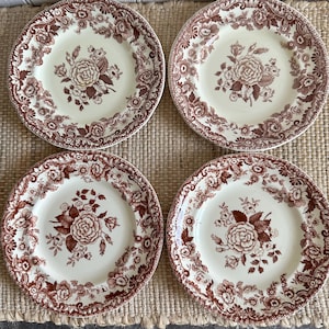 May include: Four cream-colored plates with a brown floral pattern. Each plate features a central rose design surrounded by smaller flowers and a decorative border. The plates are arranged on a woven surface.
