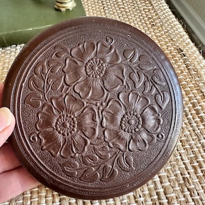 May include: A round, dark brown decorative object with a floral design. The object features three large flowers and leaves in a raised relief pattern. The object is held in a hand and appears to be made of a hard material.