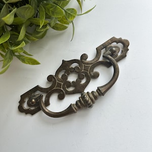 May include: A dark bronze-toned metal drawer pull featuring an ornate, elongated backplate with intricate scrollwork and cut-out details. The hardware includes a curved, swinging bail handle with a ribbed center section, attached to the backplate.