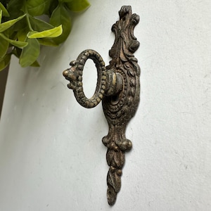 May include: A wall-mounted metal hook with a dark, weathered bronze finish. The vertical backplate features an ornate, symmetrical relief pattern with leaf-like details. A circular, textured ring extends outward from the center of the plate.