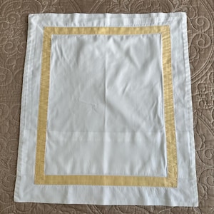May include: A rectangular white fabric placemat or napkin featuring a wide white border and a contrasting inner rectangular band in a soft pastel yellow color. The textile has a smooth, flat surface with visible stitching along the inner yellow border.