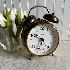 May include: A vintage-style brass twin-bell alarm clock with a white face, black Arabic numerals, and the brand name "Jerger" printed in black. The clock features black hour and minute hands with luminous tips, a red second hand, and quartz movement.