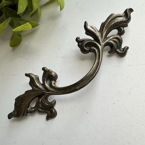 May include: A curved metal drawer pull with an antiqued bronze finish, featuring ornate, symmetrical leaf-like scrollwork at both ends. The handle has a dark, weathered patina with lighter highlights along the raised edges of the metal design.
