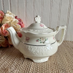 May include: A cream-colored ceramic teapot featuring a delicate floral pattern of pink and white roses. The teapot has a matching lid with a finial, a curved handle, a spout, and a silver-toned trim accenting the body and lid.