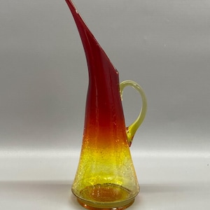 May include: A tall, decorative glass pitcher with a long, curved spout. The pitcher features a gradient of colors, transitioning from red at the top to yellow at the base. It has a yellow handle and a crackled glass texture.