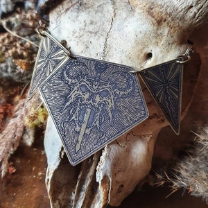 Może przedstawiać: Trzy złote metalowe zawieszki z misternymi wzorami. Zawieszka środkowa przedstawia szczegółową ilustrację czaszki z rogami i mieczem. Dwie pozostałe zawieszki mają wzór gwiazdy.