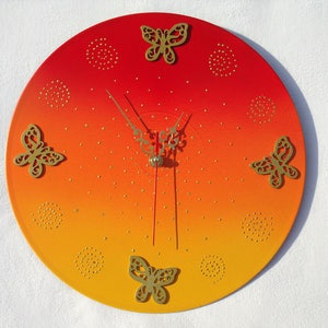 May include: A round wall clock with a gradient of red to yellow. Gold-colored butterfly cutouts and small dots decorate the clock face. The clock hands are gold-colored.