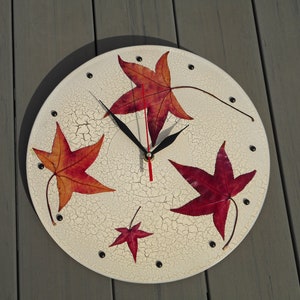 Peut inclure: Une horloge murale ronde avec un cadran crème à effet craquelé. Le cadran est orné de motifs de feuilles d'érable rouges et oranges. Des points noirs marquent les heures, et les aiguilles sont noires avec une trotteuse rouge.