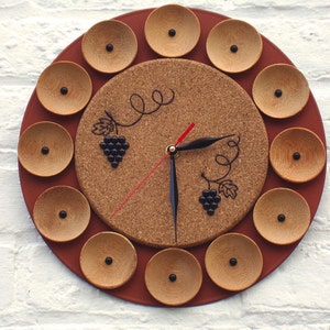 May include: A round wall clock with a cork center and a dark red border. The clock features wooden circles around the edge and black clock hands. The clock face has a grape vine design and a red second hand.