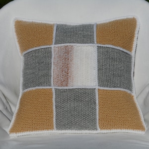 Peut inclure: Coussin carré tricoté avec un motif patchwork. Le coussin présente des carrés gris, jaune moutarde, blancs et un dégradé de marron à blanc. Le coussin est bordé de fil blanc.
