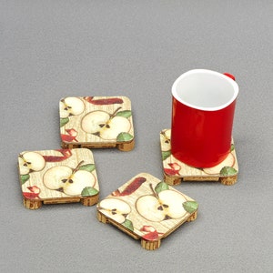 May include: Four square wooden coasters with an apple design. The coasters have a light wood grain background with apple slices, leaves, and red accents. A red mug with a white interior sits on one of the coasters.