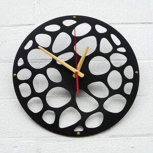May include: A black, circular wall clock with a distinctive open design. The clock face has a pattern of irregular, interconnected shapes. It has gold-coloured hands and a red second hand. The clock is mounted on a white brick wall.