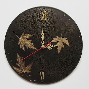 Peut inclure: Une horloge murale ronde noire avec des chiffres romains et des aiguilles dorées. Le cadran de l'horloge présente des motifs de feuilles d'érable dorées et des étoiles. L'aiguille des secondes est rouge.