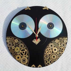 May include: A unique owl-shaped clock made from a black vinyl record. The eyes are CDs, and the clock hands are beige. The clock is decorated with gold-colored gears and small brass accents. A creative and decorative wall clock.
