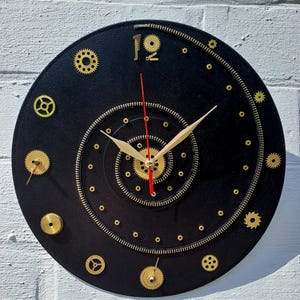 May include: A black, round clock with gold-colored gears and a zipper design. The clock hands are gold, and the second hand is red. The clock is mounted on a white brick wall. The number 12 is represented by the number 19.