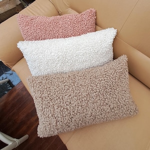 Op de afbeelding: Vier rechthoekige sierkussens in roze, wit en bruin. De kussens hebben een getextureerd, fuzzy oppervlak en zijn gerangschikt op een bruine leren bank. Decoratieve woonaccessoires.