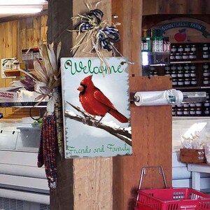 May include: A wooden sign with a red cardinal bird perched on a branch. The sign says "Welcome Friends and Family" with a green wreath above the text. The sign is hanging on a wooden post in a store.