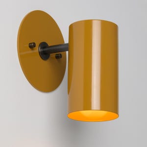 May include: A mustard yellow wall sconce with a cylindrical shade and a round back plate. The sconce has a black metal arm and is mounted on a white wall.