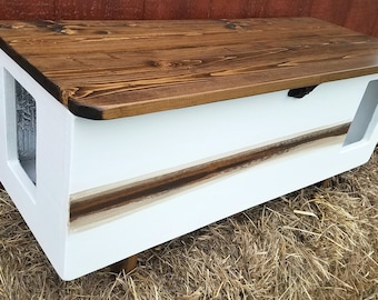 May include: A wooden cat house with a stained brown top and white body. The front and side have openings. A decorative brown stripe runs along the bottom of the white body. The cat house sits on short wooden legs.