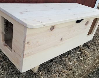 May include: A light-colored wooden cat house with a sloped roof. The house has an entrance on one side and a smaller opening on the other. The structure is elevated on small wooden legs.