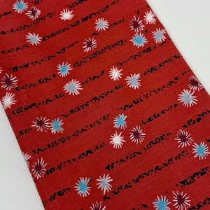 May include: A folded piece of red fabric featuring a repeating pattern of small, star-like floral motifs in white, light blue, and dark purple. The design includes thin, horizontal, wavy black lines that run across the fabric surface.
