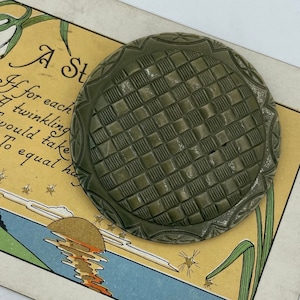 Puede incluir: Un botón de cerámica verde con un patrón tejido y un borde elevado. El botón tiene un pequeño agujero en el centro.