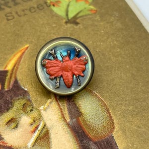 以下が含まれることがあります： 金属製の縁取りのある丸いガラスボタン。ボタンには、黒のアクセントが付いたオレンジ色の蝶の立体的なデザインが施されています。ボタンの背景は青と緑のグラデーションです。