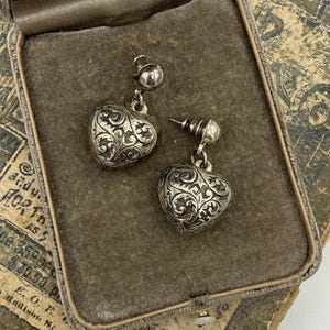 Puede incluir: Un par de pendientes plateados en forma de corazón con intrincados diseños de volutas, exhibidos en un joyero de terciopelo gris vintage. Los pendientes tienen un diseño de poste y bola. La caja descansa sobre papel envejecido con texto descolorido.