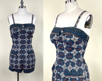 May include: A vintage romper with a blue, white, and brown geometric pattern. The romper features a square neckline, thin straps, and a fitted bodice. The shorts have pockets and a decorative border. The garment is displayed on a mannequin.