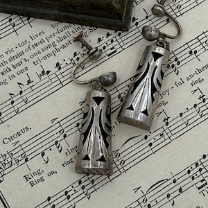May include: A pair of vintage silver-tone earrings with a geometric design and black accents. The earrings are clip-on style and rest on a sheet of vintage sheet music. A dark, square object is in the background.
