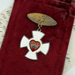 Puede incluir: Una medalla vintage en forma de cruz blanca con un emblema de corazón y corona roja, sujeta a un alfiler dorado con una estrella. La medalla descansa sobre una bolsa de terciopelo rojo oscuro, con un documento manuscrito visible.