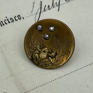 May include: A gold-toned button with a frog design and three rhinestones. The frog is sitting on a leaf with a floral design.