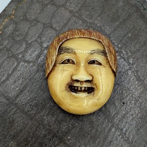 May include: A small, oval-shaped, antique mask with a smiling face. The mask is a light beige color with brown hair and detailed facial features, including open eyes and a visible row of teeth. The mask is set on a dark gray, textured surface.
