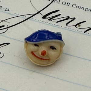 May include: A small, vintage clown face button with a blue hat, red nose, and a smiling red mouth. The button is cream-colored and has black eyes. The background is a lined paper with cursive writing.