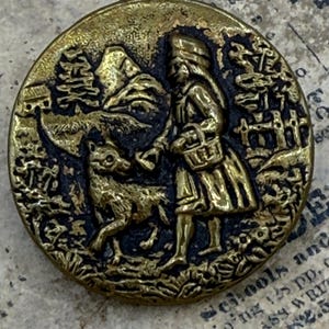 May include: An antique brass button with a detailed relief scene. The design shows a figure with a basket, a wolf, and a forest setting. The button has a dark, aged patina, and is likely made of metal.