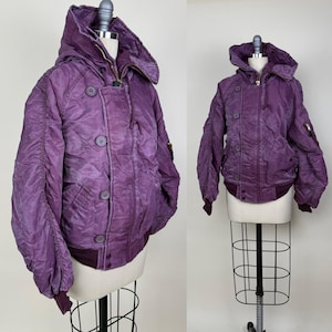 Könnte beinhalten: Eine violette Bomberjacke mit Kapuze, Knopfverschluss und gerippten Bündchen und Saum. Die Jacke hat ein gestepptes Design und zwei Vordertaschen. Die Jacke wird auf einer Schaufensterpuppe präsentiert.