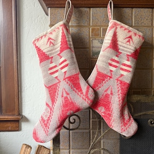 May include: Two Christmas stockings with a red and cream geometric pattern hang from a fireplace. The stockings have a loop for hanging and a traditional shape. The pattern includes diamond and stripe designs.