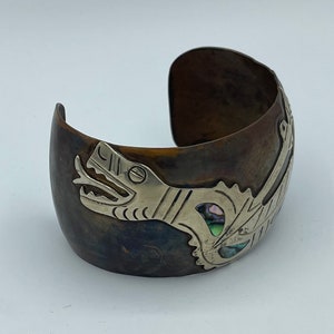 May include: A silver and copper bracelet with a stylized dragon design. The dragon is made of silver and has a turquoise and abalone shell inlay. The bracelet is open and has a slightly curved shape.