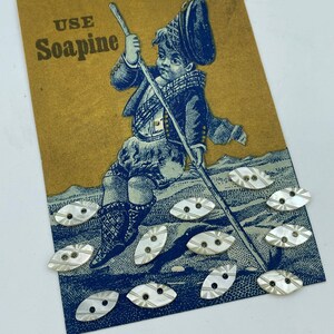 Può includere: Biglietto vintage con un'illustrazione blu di un ragazzo in abito scozzese tradizionale che tiene un bastone, con la scritta "USE Soapine" in oro. Bottoni ovali in madreperla con un design a righe e due fori sono sparsi sulla carta.