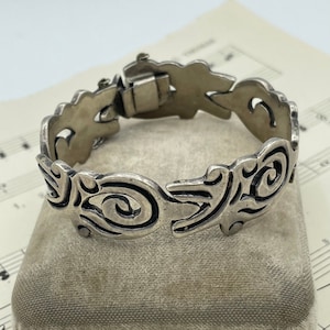 May include: A silver-toned bracelet with an openwork design featuring swirling patterns. The bracelet is resting on a small, gray velvet box. The clasp is visible, and the bracelet appears to be vintage or antique.