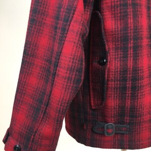 May include: A close-up of a red and black plaid jacket. The jacket features a buttoned sleeve, a pocket, and a buckle detail at the waist. The fabric appears to be a thick wool or wool blend.
