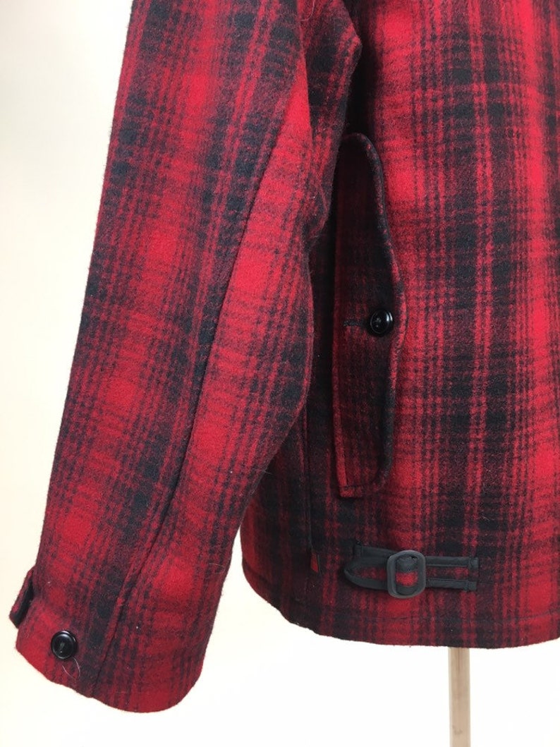 May include: A close-up of a red and black plaid jacket. The jacket features a buttoned sleeve, a pocket, and a buckle detail at the waist. The fabric appears to be a thick wool or wool blend.