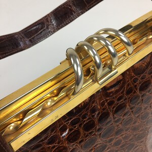May include: A brown crocodile skin handbag with a gold and silver clasp. The clasp has a twisted design and multiple loops.