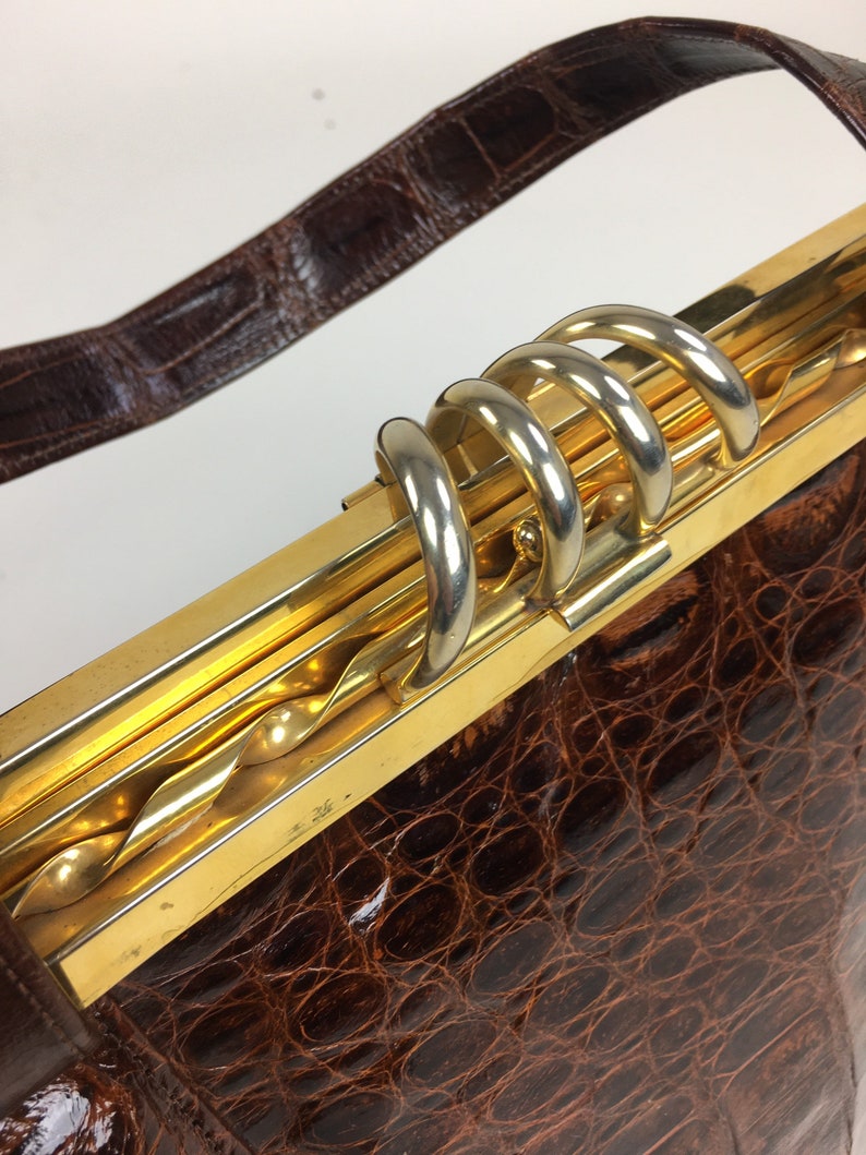 May include: A brown crocodile skin handbag with a gold and silver clasp. The clasp has a twisted design and multiple loops.