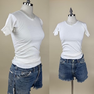 May include: Two images of a white, short-sleeved t-shirt with a scoop neck and distressed denim shorts. The t-shirt is form-fitting and has a slightly textured appearance. The denim shorts are blue with a frayed hem.