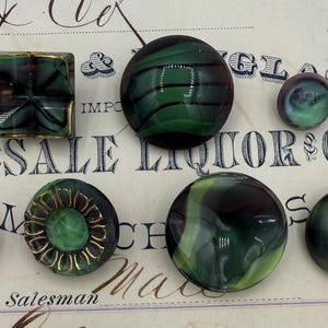 May include: A collection of vintage green and black glass buttons in various shapes, including round, square, and floral designs. Some buttons have gold accents. The buttons are displayed on a vintage paper background with faded text.