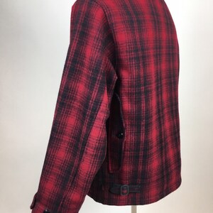 May include: A red and black plaid jacket with a button closure and a small buckle detail at the waist. The jacket has a classic collar and long sleeves. The fabric appears to be a thick wool or wool blend.